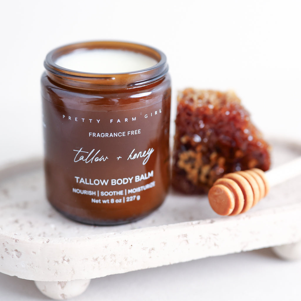 Pretty Farm Girl Tallow + Honey Tallow Body Balm jar on a white stone surface with honey dipper and honeycomb.
