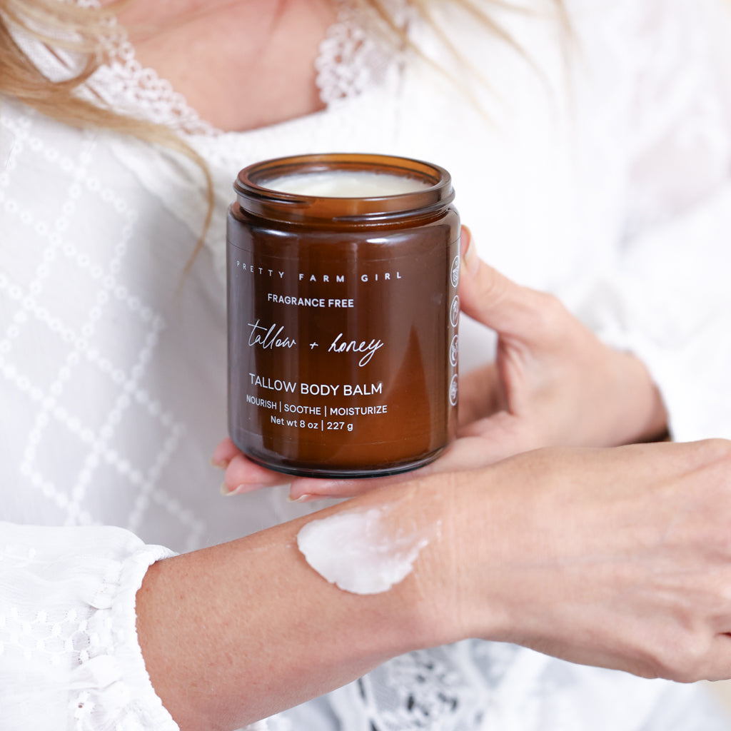 Pretty Farm Girl Tallow + Honey Tallow Body Balm jar held in hand on a soft white fabric background.