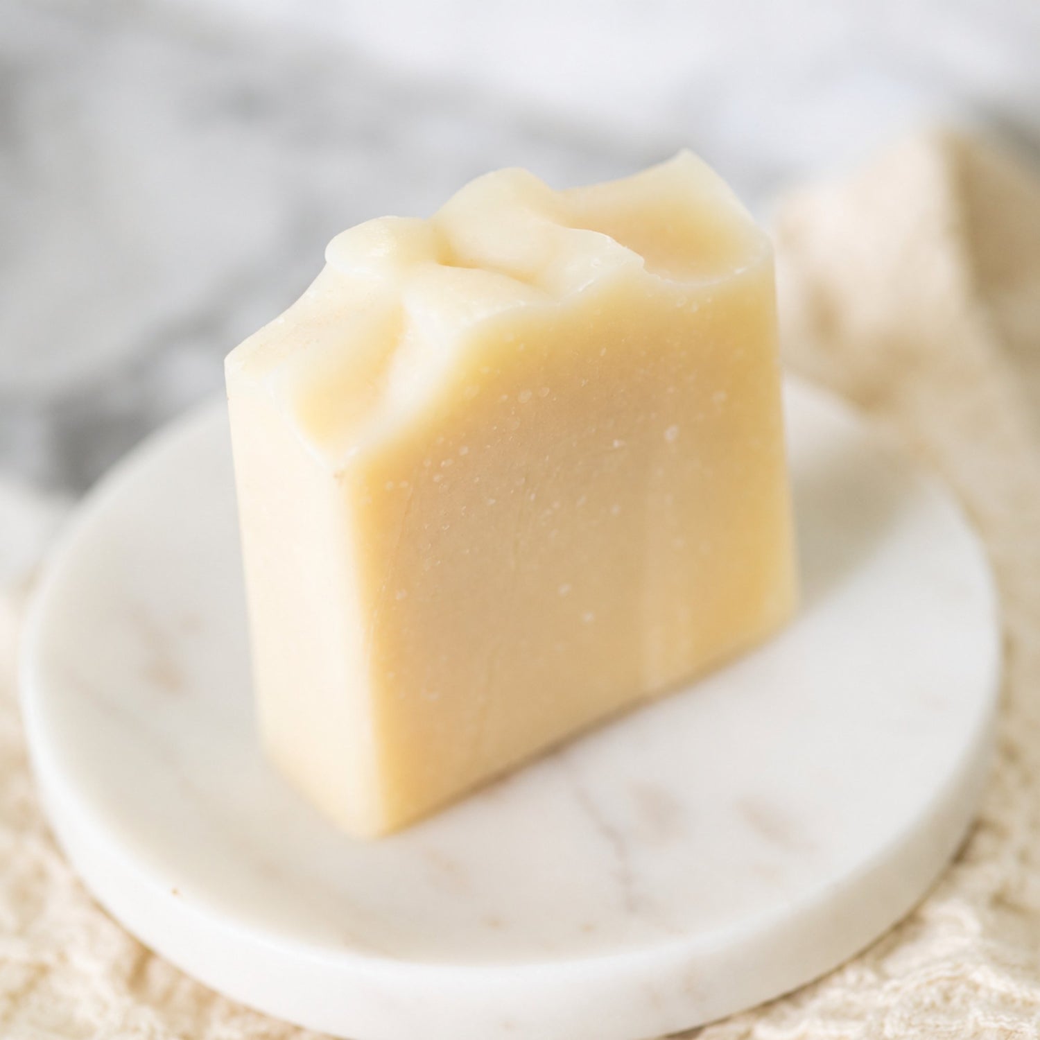 A white bar of handmade tallow soap with a textured, organic top surface, resting on a small white marble dish over a beige fabric surface.