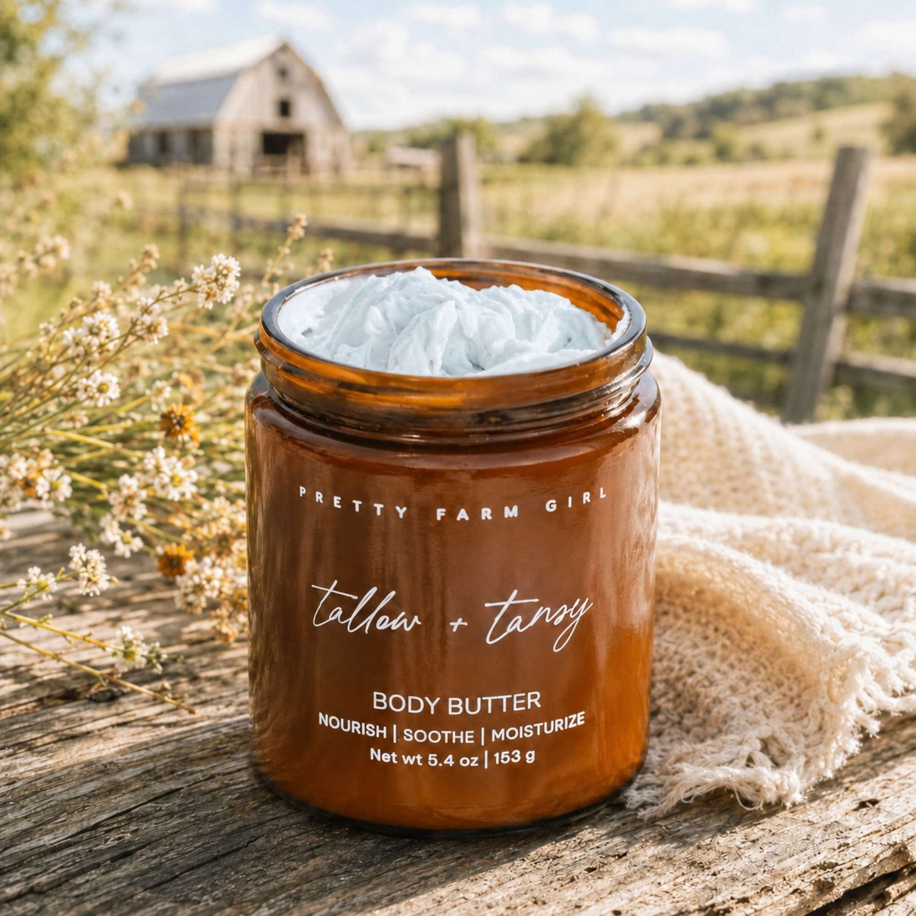 An amber glass jar of Pretty Farm Girl 'tallow + tansy' body butter sits on a rustic wooden surface outdoors, with a white body butter cream visible at the top and a beige woven cloth beside it. A blurred farm background with a barn, fence, and greenery is visible.