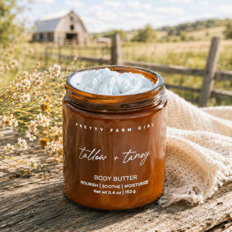 An amber glass jar of Pretty Farm Girl 'tallow + tansy' body butter sits on a rustic wooden surface outdoors, with a white body butter cream visible at the top and a beige woven cloth beside it. A blurred farm background with a barn, fence, and greenery is visible.