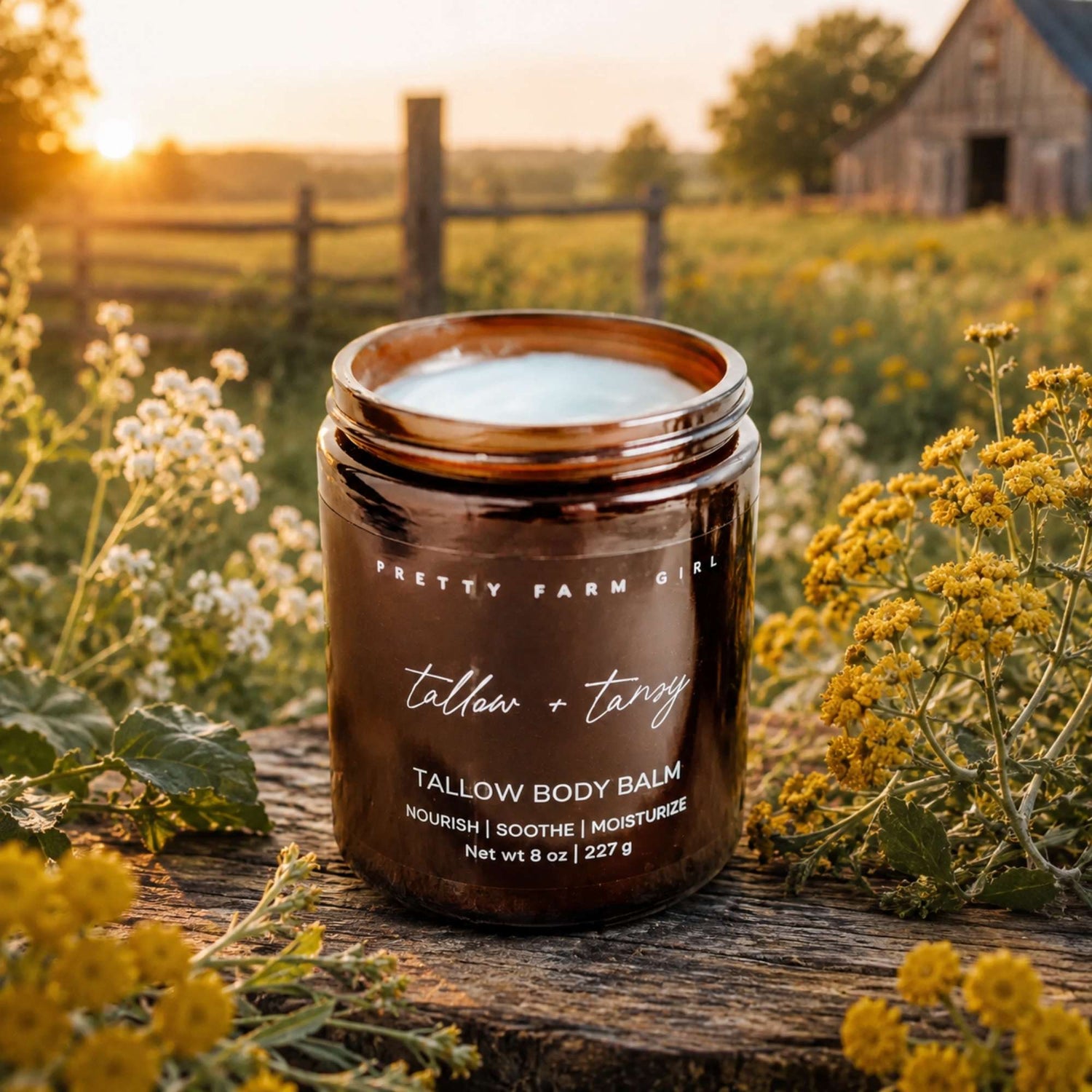 A jar of Pretty Farm Girl Tallow Body Balm sits on a rustic wooden fence in a sunlit field, surrounded by white and yellow wildflowers, with a barn and trees in the background.