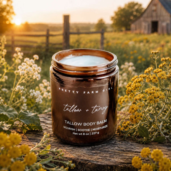 A jar of Pretty Farm Girl Tallow Body Balm sits on a rustic wooden fence in a sunlit field, surrounded by white and yellow wildflowers, with a barn and trees in the background.