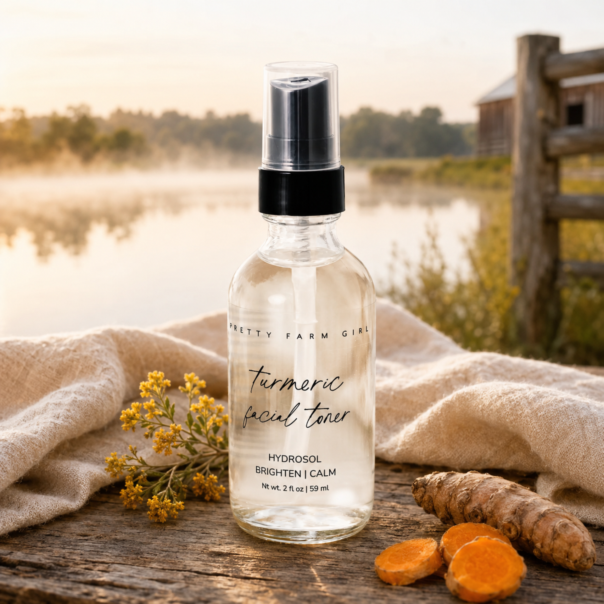 A clear glass spray bottle labeled Pretty Farm Girl Turmeric Facial Toner stands on a rustic wooden surface outdoors, with a beige cloth, small yellow flowers, turmeric root, and turmeric slices around it. A blurred lake, trees, and wooden fence are in the background.