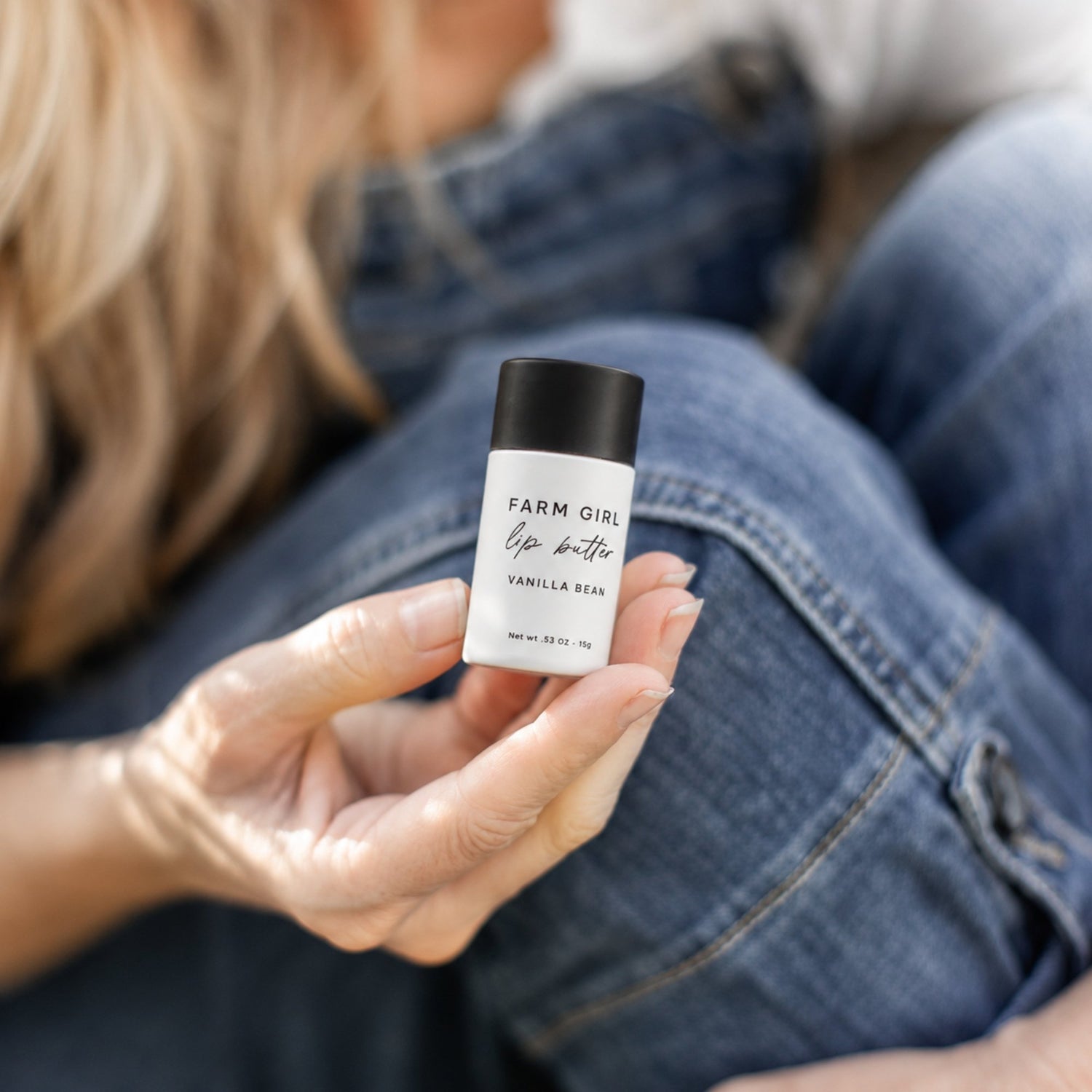 A person in a denim jacket and jeans holds a small white and black stick of Farm Girl Lip Butter in Vanilla Bean scent.