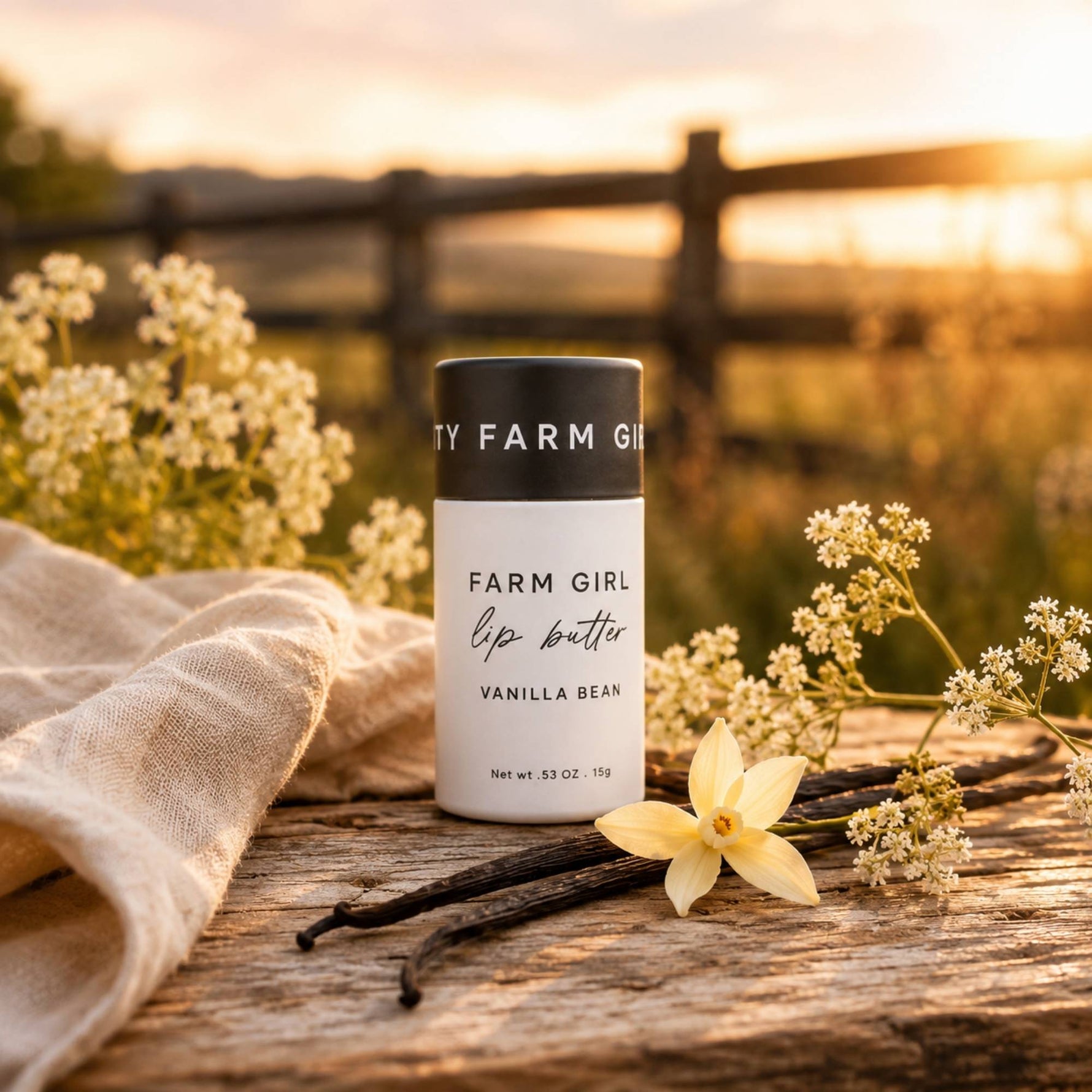 A white and black lip balm container labeled 'FARM GIRL lip butter VANILLA BEAN' stands on a rustic wooden surface outdoors at sunset, surrounded by cream flowers, vanilla beans, and a beige cloth.
