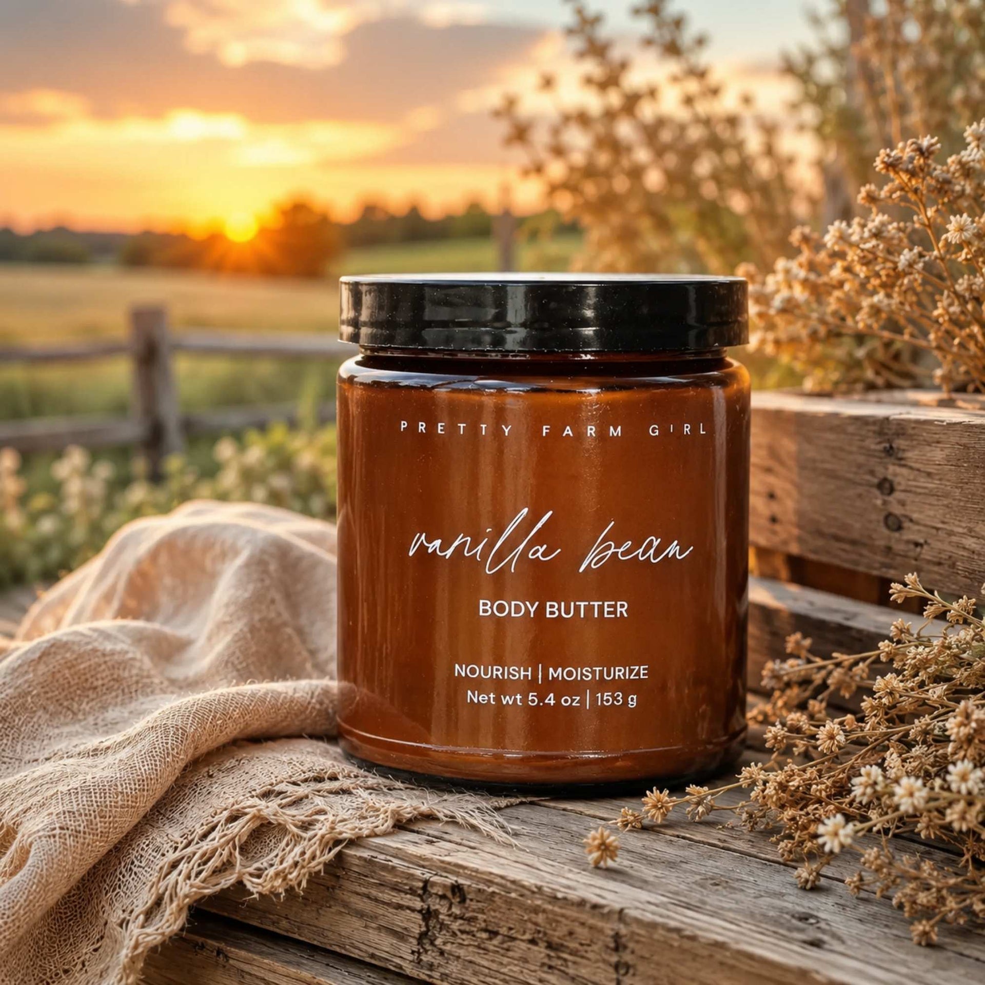 A jar of Pretty Farm Girl 'Vanilla Bean' body butter sits on a rustic wooden crate draped with a beige linen cloth, surrounded by dried flowers, with a blurred sunset field and fence in the background.