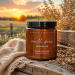 A jar of Pretty Farm Girl 'Vanilla Bean' body butter sits on a rustic wooden crate draped with a beige linen cloth, surrounded by dried flowers, with a blurred sunset field and fence in the background.