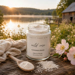A glass jar of 'PRETTY FARM GIRL wild rose CLEANSING GRAINS' sits on a rustic wooden table outdoors by a lake at sunset, with a cloth, spoon, and pink flowers around it.