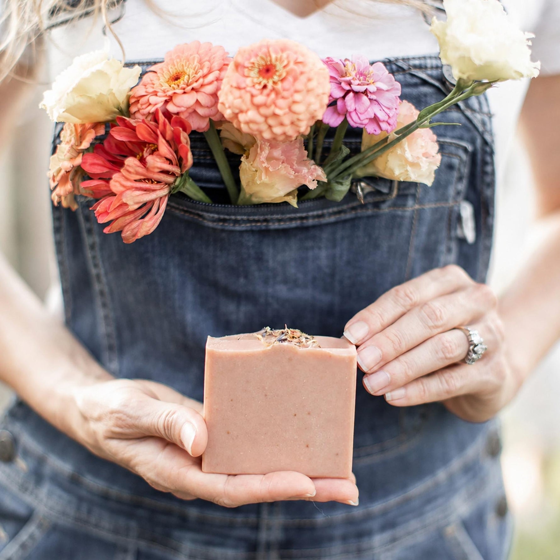 A person wearing blue denim overalls holds a Wild Rose + Honey soap bar in their hands, with a bouquet of colorful flowers tucked into the front pocket of the overalls.