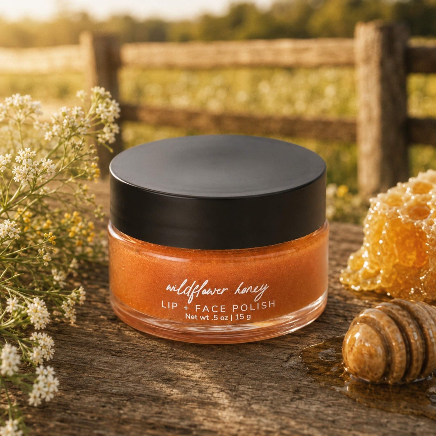 A jar of Wildflower Honey Lip + Face Polish sits on a rustic wooden surface outdoors, surrounded by wildflowers, a honeycomb piece, and a wooden honey dipper. The product is in a clear glass jar showing a warm amber-orange polish, with a matte black lid.