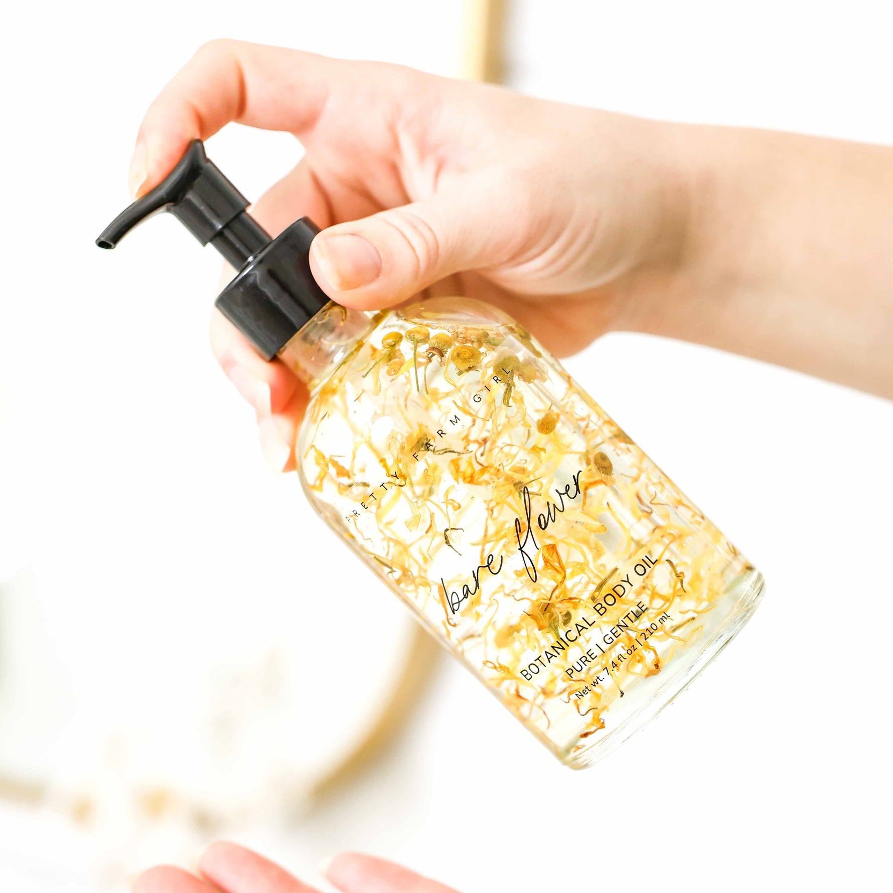 Hand holding a bottle of 'Bare Flower' botanical body oil with visible flower petals in the bottle.