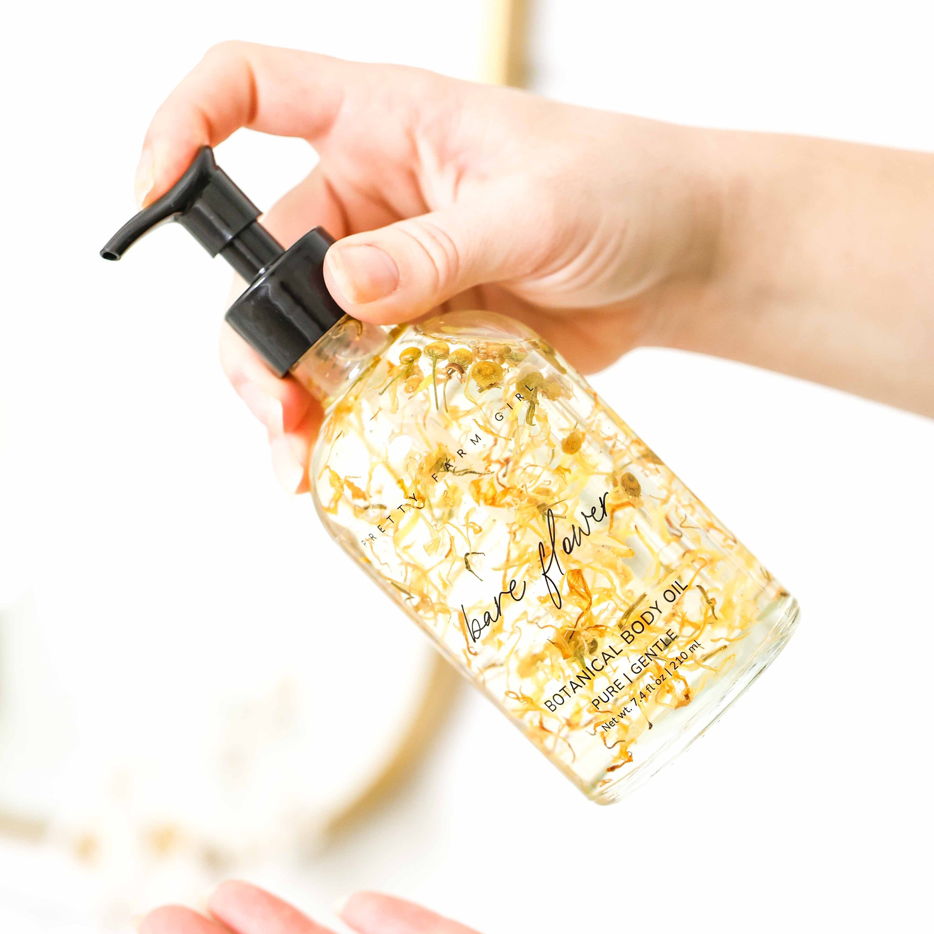 Hand holding a bottle of 'Bare Flower' botanical body oil with visible flower petals in the bottle.