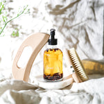 Bottle of 'lymph love' botanical body oil on a white surface with a wooden dry brush, bamboo boody gua sha, and a plant in the background.