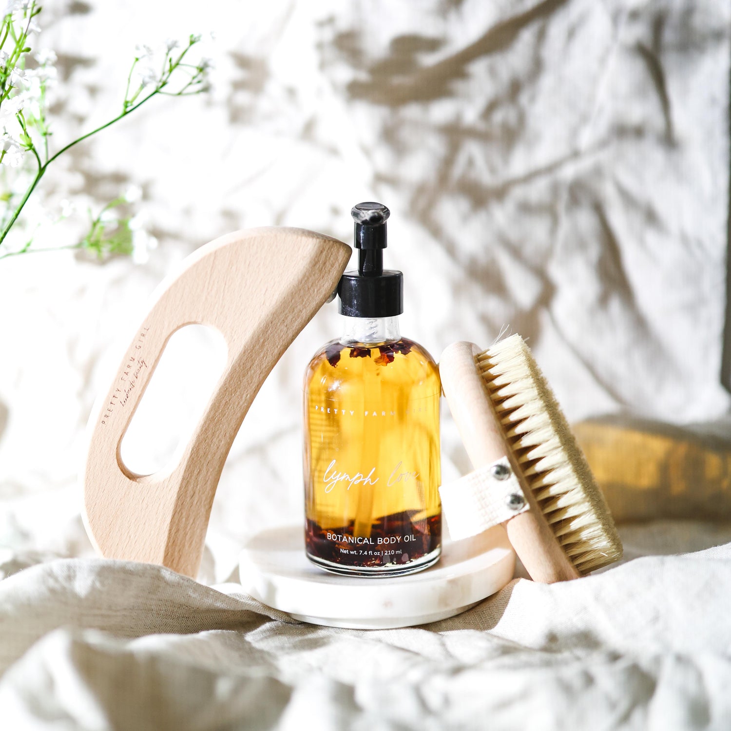 Bottle of 'lymph love' botanical body oil on a white surface with a wooden dry brush, bamboo boody gua sha, and a plant in the background.