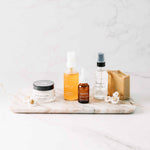 Collection of skincare products, the fresh face tallow cream, farm girl cleansing oil, brightening vitamin c serum, turmeric toner, and brightening soap bar on a marble tray with a white background