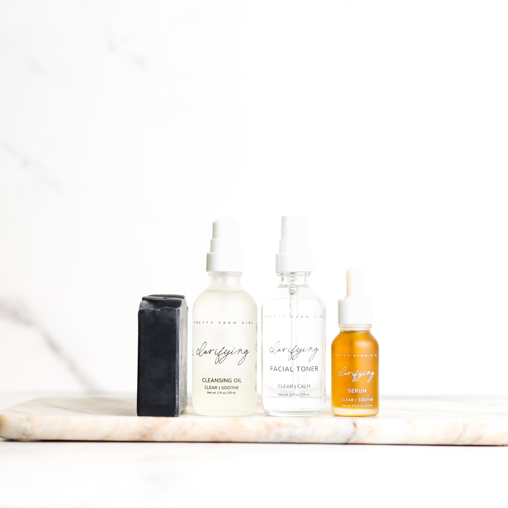 Set of skincare products including a bar of soap, clarifying toner and cleansing oil bottles, and a serum on a marble surface with a white background.