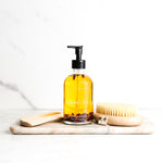 Bottle of body oil with a dry brush and wooden gua sha on a marble slab against a white background