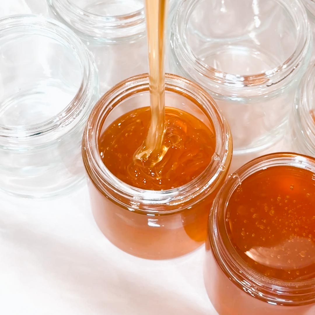 Honey being poured into a glass jar with other jars around on a white background