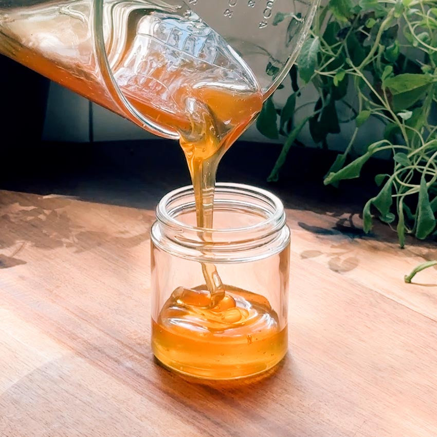 Honey being poured from a jar into another jar on a wooden surface with plants in the background.