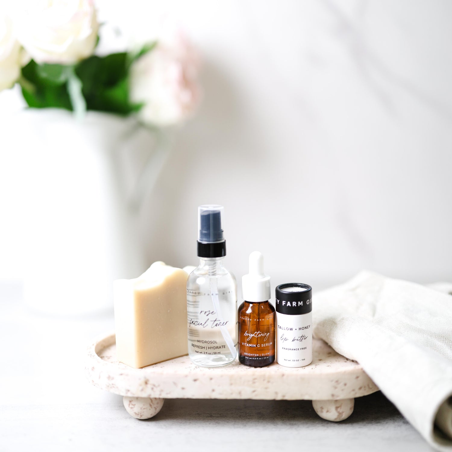 Pretty Farm Girl's Rooted and Ready Kit, showing 4 products including a Fresh Face Cream Soap Bar, 2oz Rose Facial Toner, .5oz Brightening Vitamin C Serum, .5oz Tallow + Honey Fragrance Free Lip Butter in an Eco-Friendly paper tube on a tray with a blurred vase in the background.