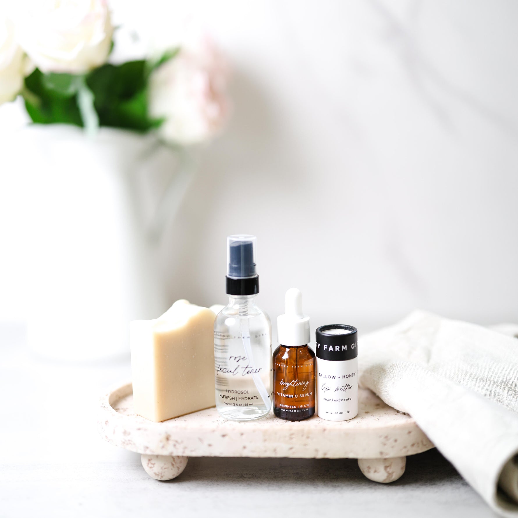 Pretty Farm Girl's Rooted and Ready Kit, showing 4 products including a Fresh Face Cream Soap Bar, 2oz Rose Facial Toner, .5oz Brightening Vitamin C Serum, .5oz Tallow + Honey Fragrance Free Lip Butter in an Eco-Friendly paper tube on a tray with a blurred vase in the background.