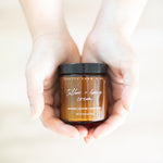 Brown jar of 'Tallow + Honey Cream' held in hands against a light background