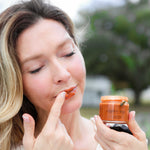 Woman applying Wildflower Honey Lip + Face Polish to her lips outdoors, holding the jar with a bee on it.