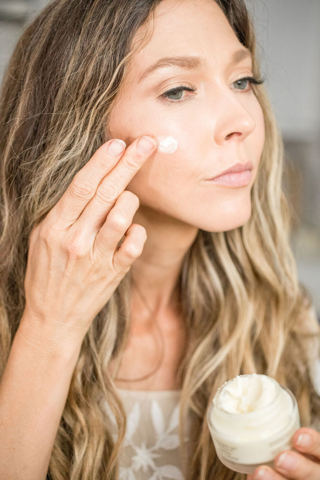 Close-up of a woman applying Pretty Farm Girl tallow face cream to her cheek with an open jar in hand